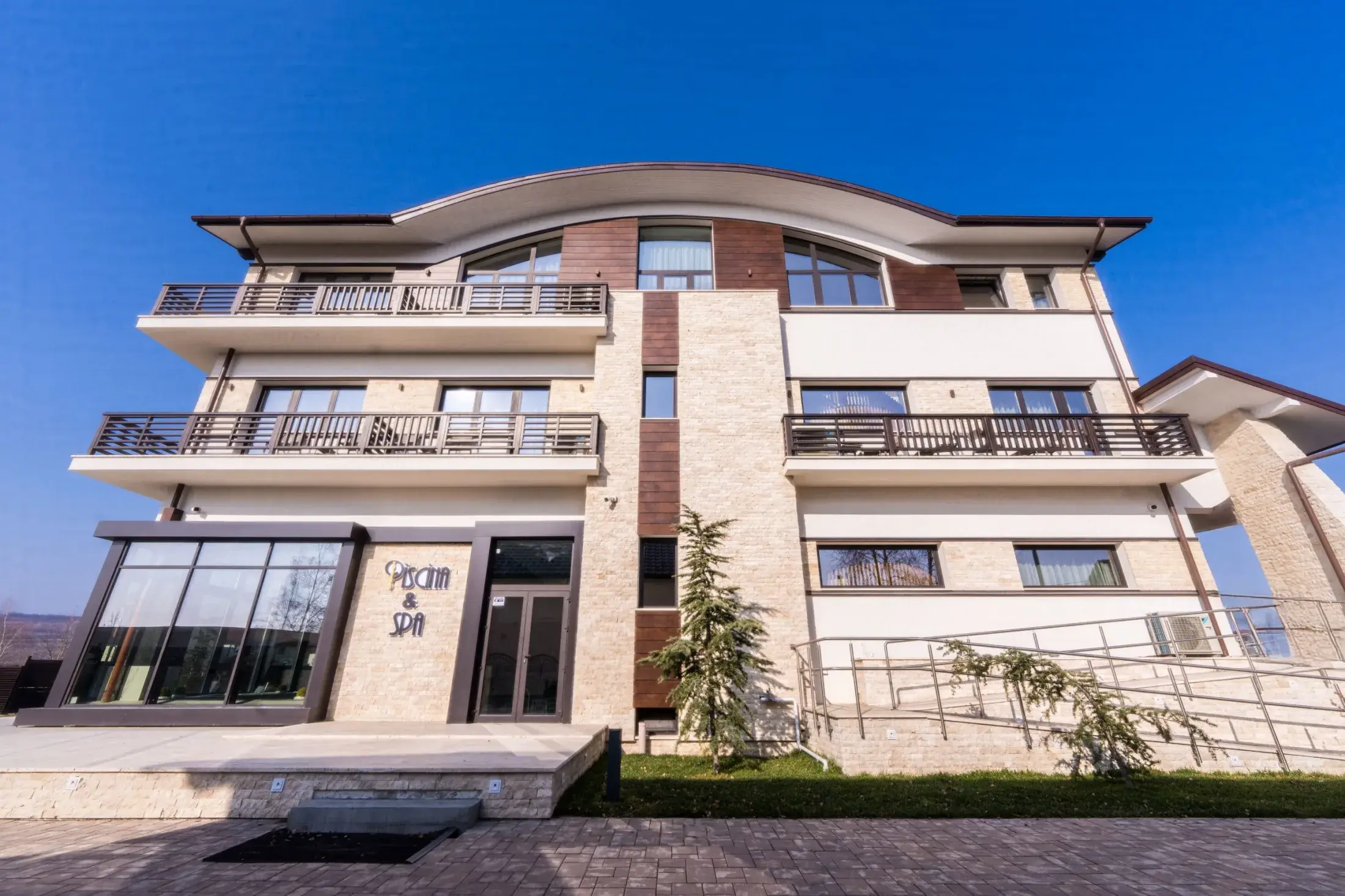 Clădirea pensiunii pe trei etaje, cu balcoane și intrarea în zona de piscină și SPA