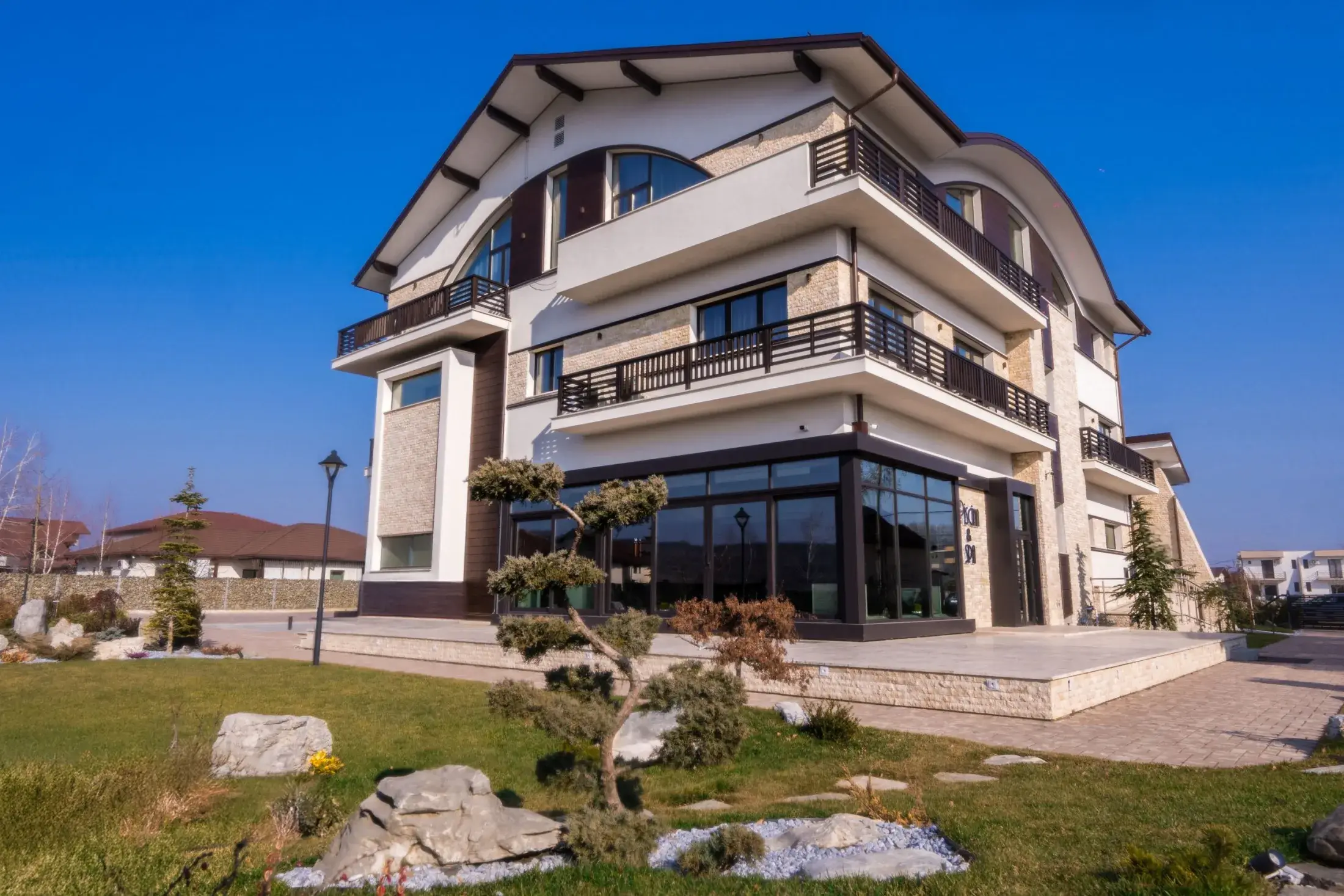 Pensiunea văzută din grădină, cu gazon, pietre mari și un brad tuns