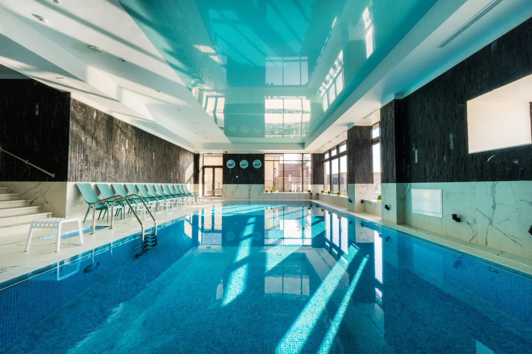 Piscina interioară, cu șezlonguri turcoaz pe margine și ferestre spre grădină